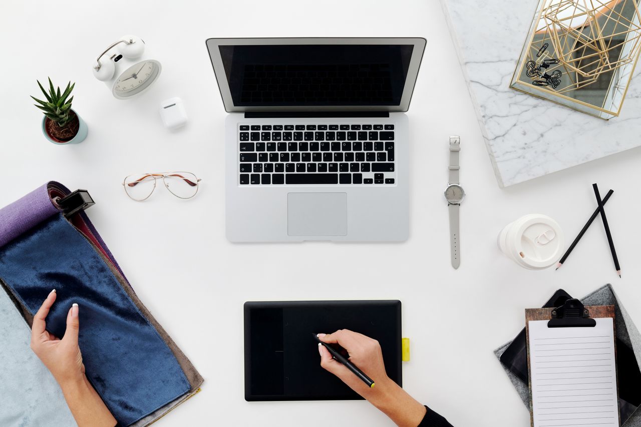 Offerings Overhead view of a designer's workspace with a laptop, drawing tablet, and office supplies on a white surface.